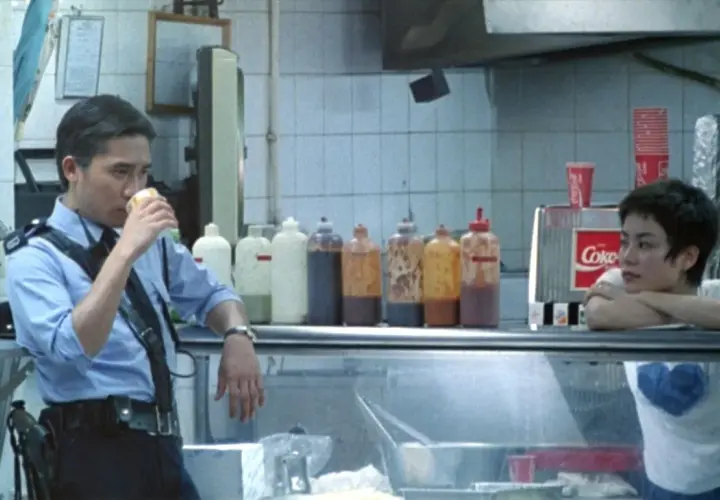 Un poliziotto sorseggia una bevanda, mentre una ragazza appoggia il mento sulle mani. Scena da "Hong Kong Express" con spiegazione implicita.