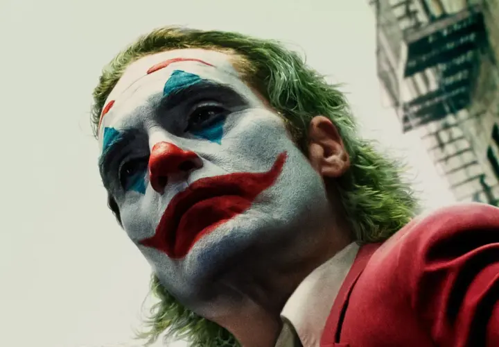 Il volto truccato di chi è Joker, con capelli verdi e un sorriso rosso dipinto, guarda verso l'alto.