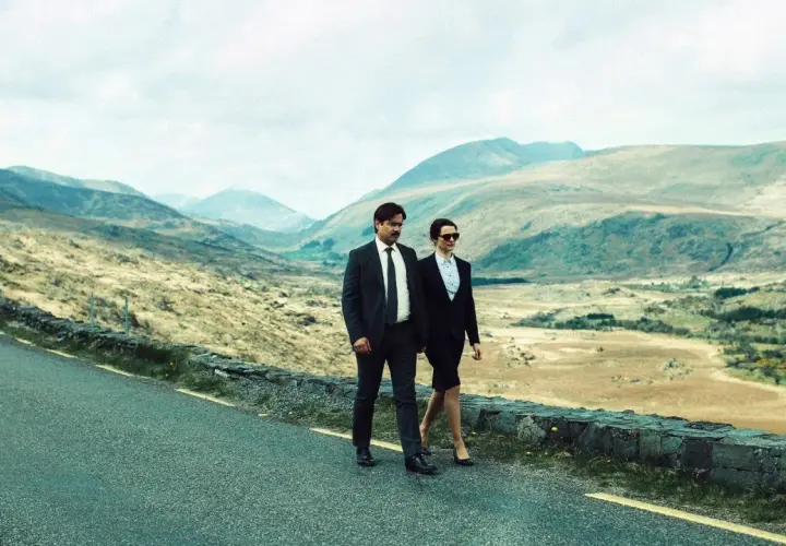 Coppia in abiti eleganti cammina su strada panoramica, evocando il significato di "The Lobster" e la ricerca di un legame in un paesaggio desolato.