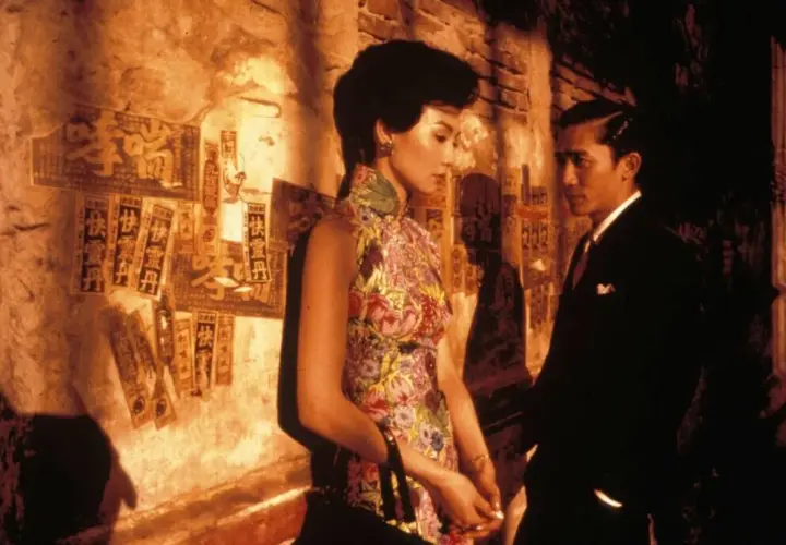 In the mood for love spiegazione: una donna in cheongsam fiorito e un uomo in abito scuro si guardano con intensità, sullo sfondo di un muro con scritte cinesi.