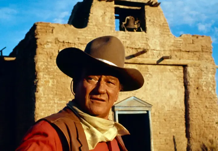 John Wayne, icona del West, con cappello e bandana, davanti a una chiesa diroccata. Ricorda le sue frasi western famose.