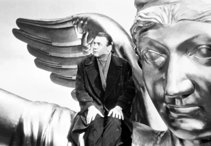 Un uomo seduto su un'ala d'angelo dorata, con un volto statuario di fronte. Un'immagine iconica per capire "il cielo sopra berlino".