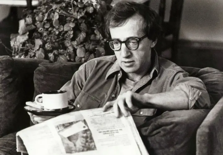 Woody Allen, con occhiali e camicia, legge un giornale con una tazza di caffè, forse pensando al suo prossimo Oscar.