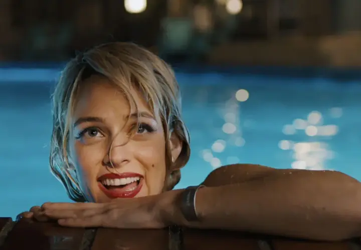 Una donna sorride dall'acqua di una piscina, con luci sfocate sullo sfondo. Una scena che ricorda "Under the Silver Lake".