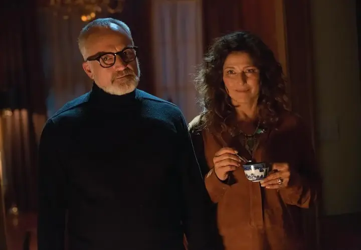 Coppia anziana in un interno buio, lui con occhiali e dolcevita nero, lei sorride con una tazza in mano. Sembra il **get out finale**.