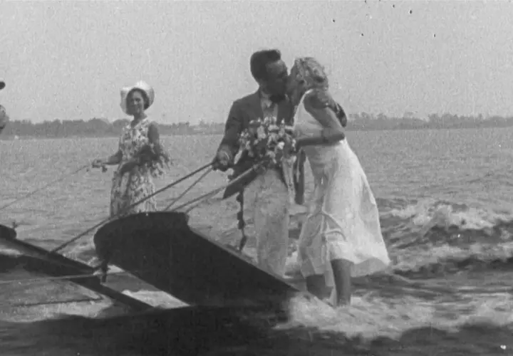 Coppia di sposi baciarsi su una barca trainata, con un uomo in cilindro e una donna con cappello che li seguono. Un'immagine che sembra uscita da un film di 100 anni fa.