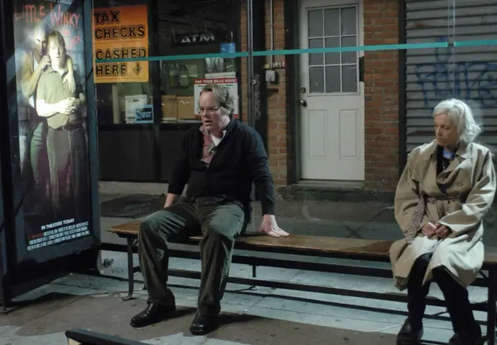 Uomo e donna seduti su una panchina, vicino a un poster di "Little Winky". Una scena che evoca la complessità di "Synecdoche, New York".