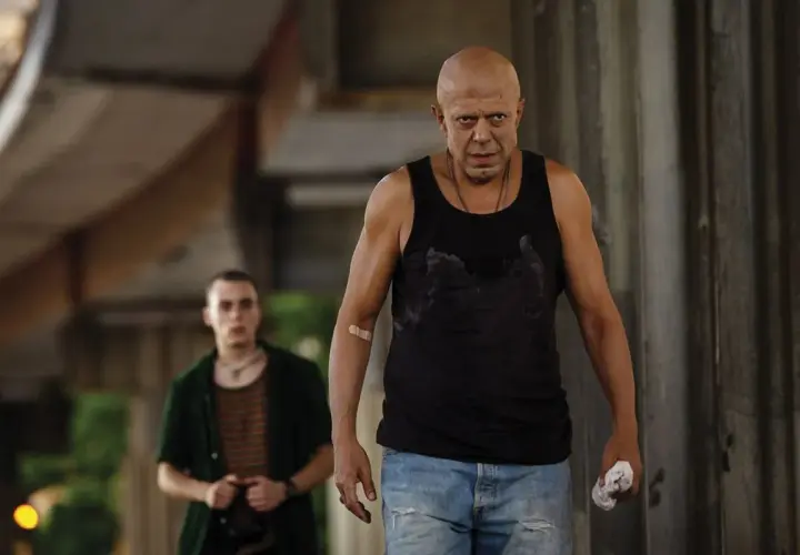 Due uomini, uno calvo e con un cerotto sul braccio, camminano in un ambiente urbano. Scena dal film "Adagio".