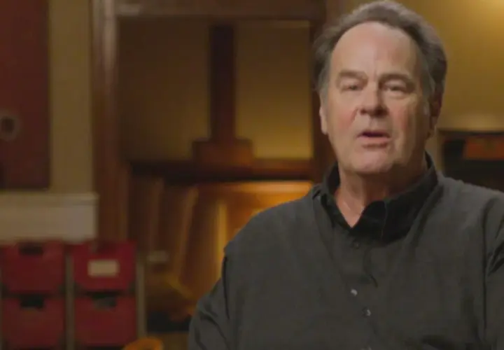 Dan Aykroyd oggi, con un'espressione pensierosa, indossa una camicia scura. Sullo sfondo, oggetti d'arte e sedie rosse.