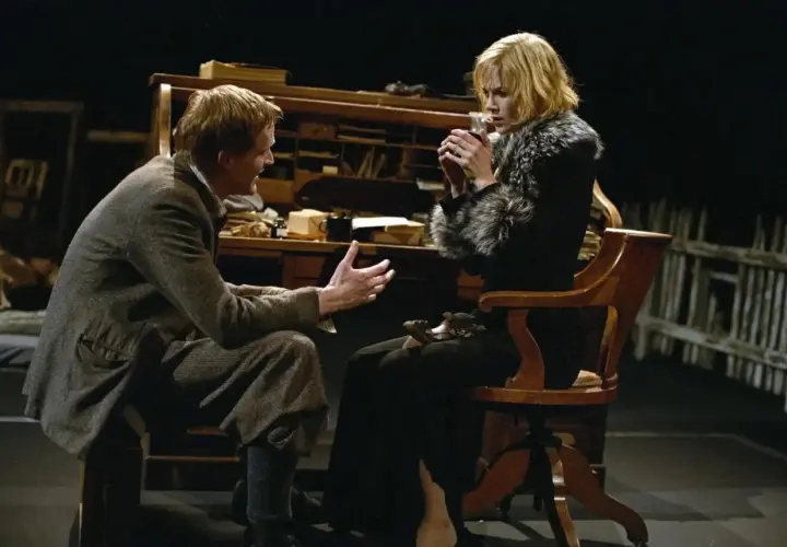 Una scena intensa da Dogville recensione: un uomo parla animatamente a una donna seduta, che tiene una tazza tra le mani.
