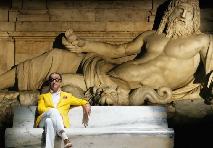 Un uomo in giacca gialla seduto su una panchina di marmo, con una statua antica alle spalle. Un'immagine che evoca la grande bellezza, spiegazione visiva dell'arte e del tempo.