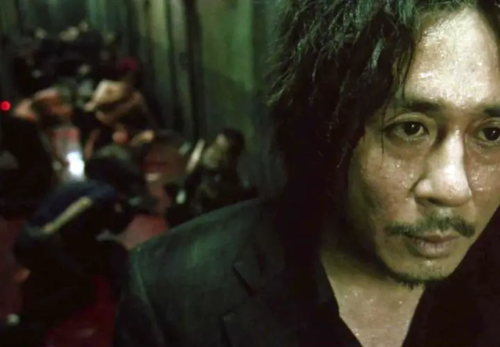 Primo piano di un uomo sudato, con un'espressione intensa, sullo sfondo una scena confusa di persone. Ricorda il film Oldboy e il suo polpo.