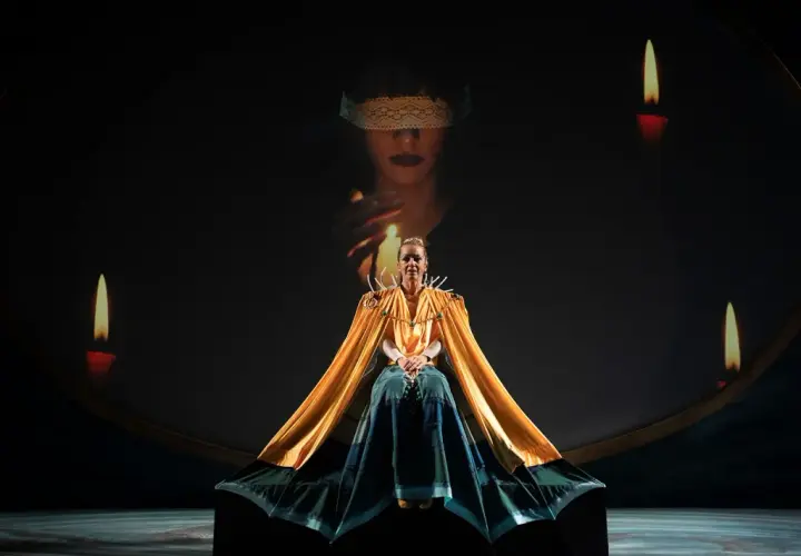 Scena teatrale ispirata al circe film: una donna con mantello dorato, circondata da candele, sotto un'immagine proiettata di una figura bendata.