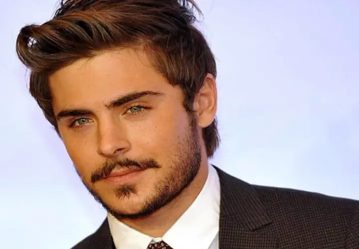 Primo piano di Zac Efron giovane, con sguardo intenso e barba curata, indossa una giacca scura.