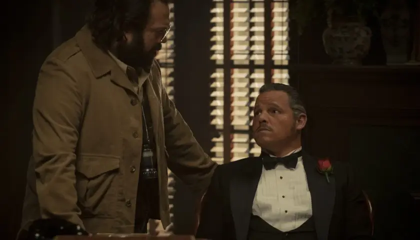 Un uomo con la barba e occhiali parla con un altro in smoking con una rosa. La scena suggerisce una recensione di un'offerta importante.