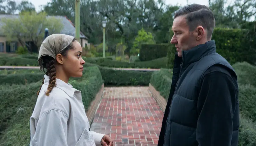 Due persone si guardano in un giardino curato, con siepi e un vialetto di mattoni. Le recensioni per "il maestro giardiniere" potrebbero descrivere scene come questa.