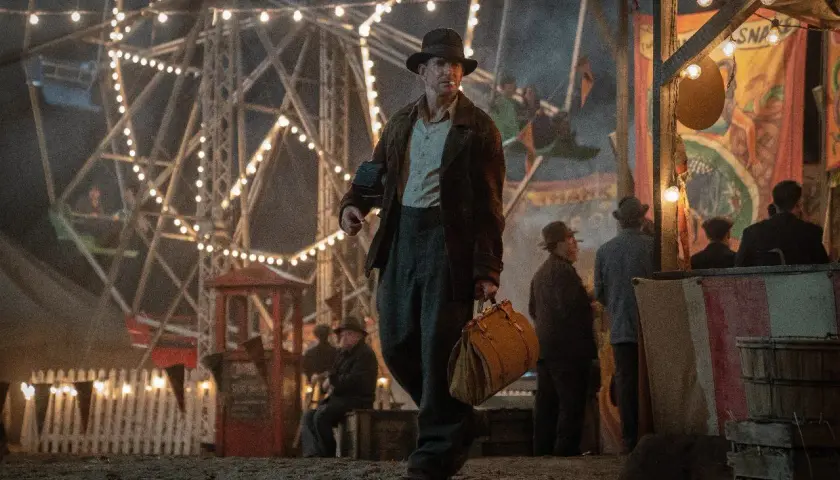Un uomo con un cappello cammina in una fiera delle illusioni, con una ruota panoramica illuminata sullo sfondo. Le recensioni di Nightmare Alley descrivono questo mondo oscuro.
