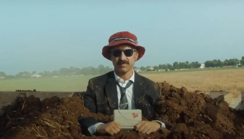 Uomo in abito e cappello rosso, sepolto nel terreno, tiene una lettera con un cuore. Una scena surreale per la recensione "il migliore dei mondi".
