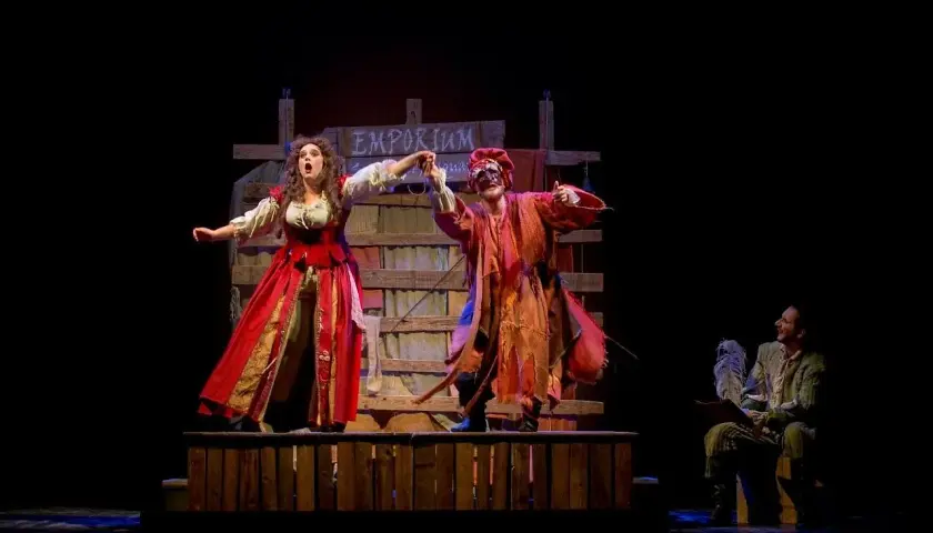Scena teatrale con due attori in costume, uno con abito rosso e l'altra con maschera e vestiti arancioni. Sullo sfondo un cartello "Emporium". Le recensioni per Romeo è Giulietta lodano queste performance.
