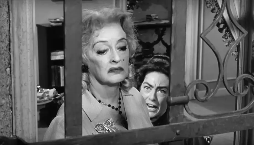 Bette Davis anziana, con un'espressione di disgusto, è dietro una grata. Un'altra donna con occhi sbarrati la spia.