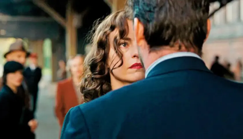 Una donna con labbra rosse e capelli ricci guarda intensamente un uomo, il segreto del suo volto trama. Sullo sfondo, persone sfocate in una stazione.