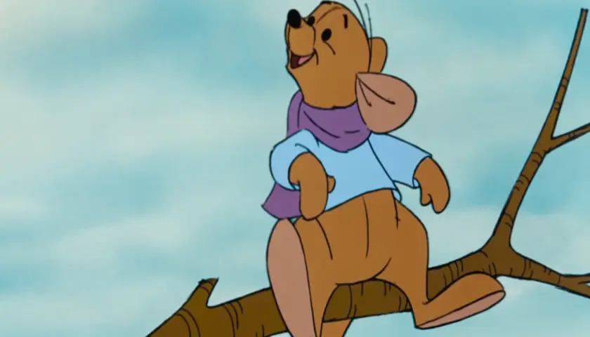 Winnie the Pooh, interpretato da Jim Cummings, siede su un ramo con una sciarpa viola e una camicia azzurra.