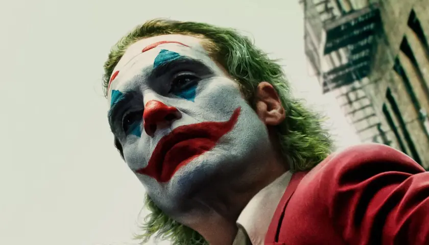 Il volto truccato di chi è Joker, con capelli verdi e un sorriso rosso dipinto, guarda verso l'alto.