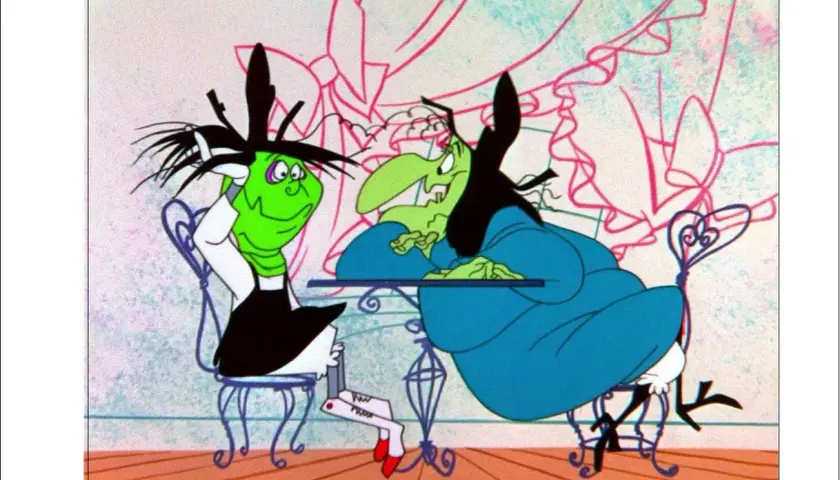Due streghe, una verde e una con la pelle verde, sedute a un tavolo. La strega Hazel, con i capelli neri e un vestito blu, sembra annoiata.