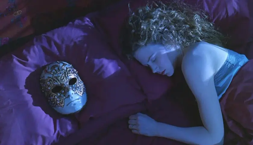 Una donna dorme su un letto di seta viola, accanto a una maschera veneziana ornata. La sua curiosità è palpabile, come se stesse sognando occhi chiusi.
