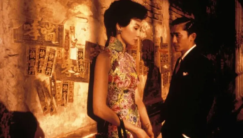 In the mood for love spiegazione: una donna in cheongsam fiorito e un uomo in abito scuro si guardano con intensità, sullo sfondo di un muro con scritte cinesi.