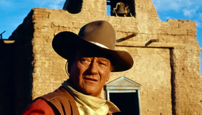 John Wayne, icona del West, con cappello e bandana, davanti a una chiesa diroccata. Ricorda le sue frasi western famose.