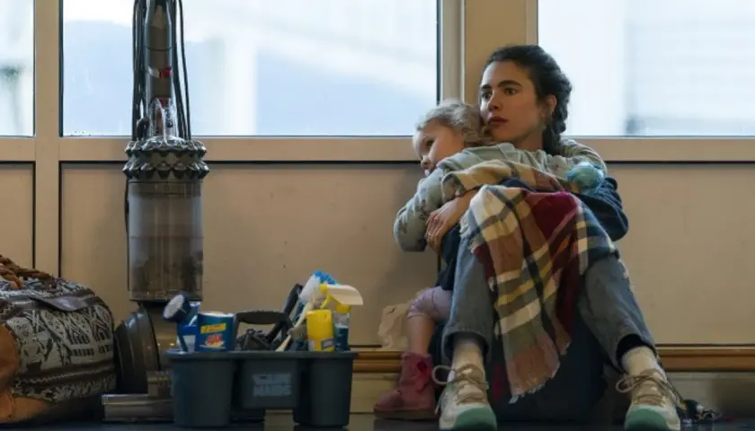 Madre abbraccia figlia, circondata da attrezzi per le pulizie e un aspirapolvere. Le recensioni per il suo lavoro di maid sono positive, ma la vita è dura.