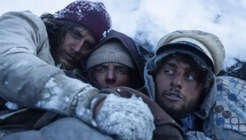 Tre uomini stremati nel gelo, un'immagine che evoca le recensioni de "La società della neve", la lotta per la sopravvivenza.
