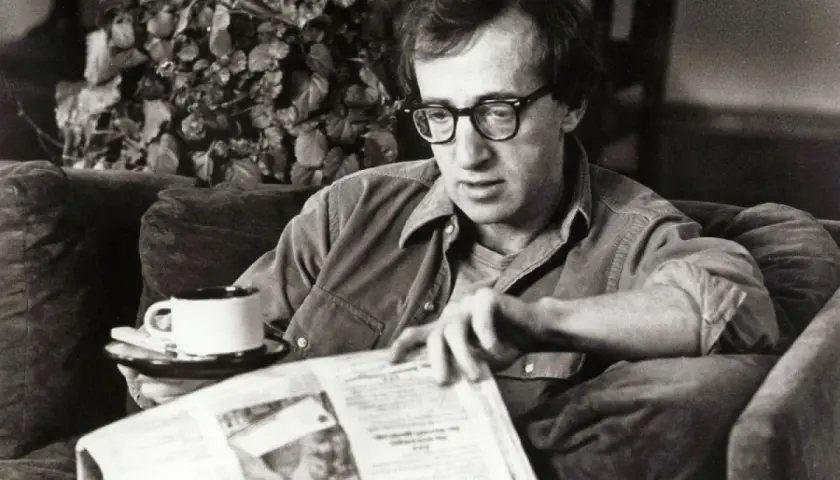Woody Allen, con occhiali e camicia, legge un giornale con una tazza di caffè, forse pensando al suo prossimo Oscar.