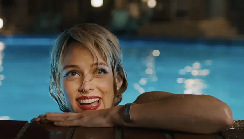 Una donna sorride dall'acqua di una piscina, con luci sfocate sullo sfondo. Una scena che ricorda "Under the Silver Lake".