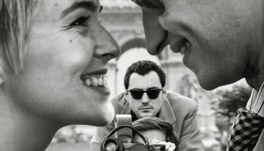 Primo piano di una donna sorridente, con un uomo che la guarda e un altro uomo con occhiali da sole che filma. Un omaggio ai registi importanti.