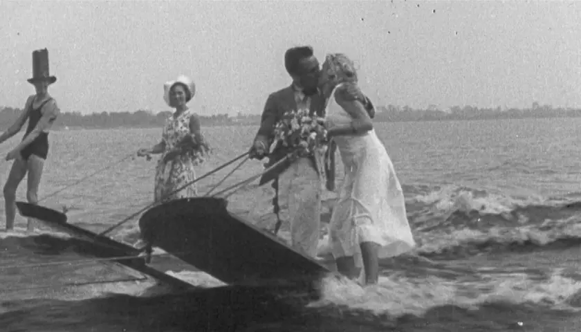 Coppia di sposi baciarsi su una barca trainata, con un uomo in cilindro e una donna con cappello che li seguono. Un'immagine che sembra uscita da un film di 100 anni fa.