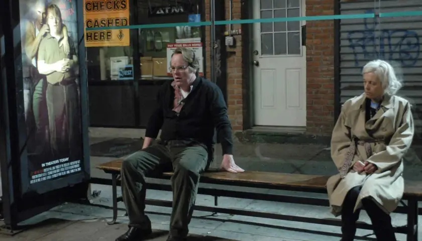 Uomo e donna seduti su una panchina, vicino a un poster di "Little Winky". Una scena che evoca la complessità di "Synecdoche, New York".