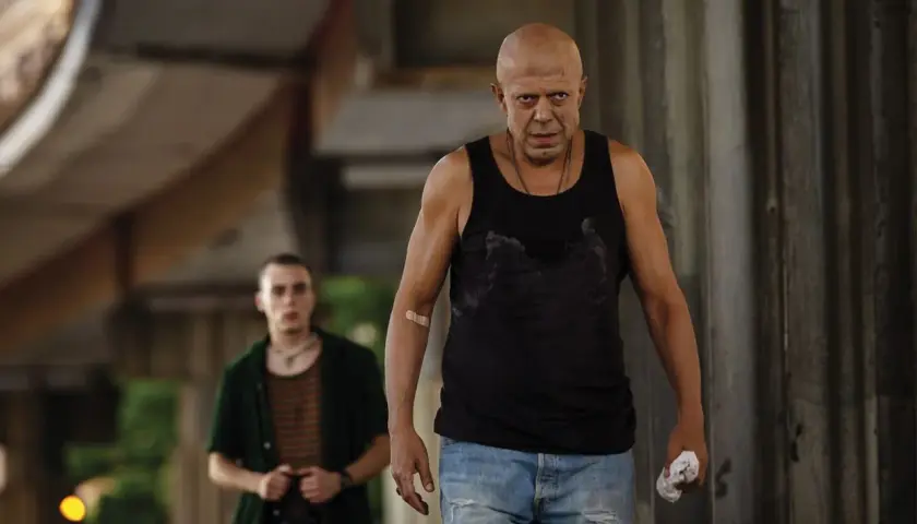 Due uomini, uno calvo e con un cerotto sul braccio, camminano in un ambiente urbano. Scena dal film "Adagio".