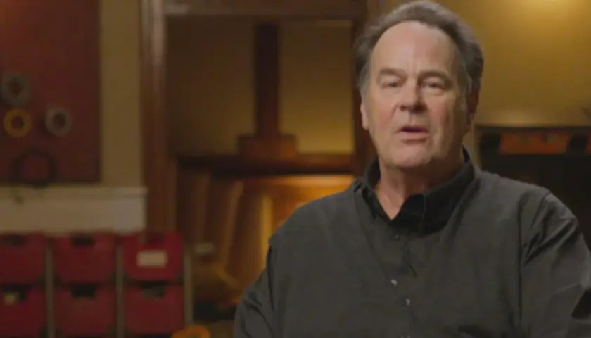 Dan Aykroyd oggi, con un'espressione pensierosa, indossa una camicia scura. Sullo sfondo, oggetti d'arte e sedie rosse.