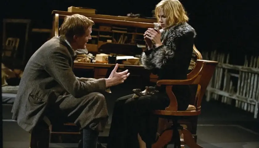 Una scena intensa da Dogville recensione: un uomo parla animatamente a una donna seduta, che tiene una tazza tra le mani.