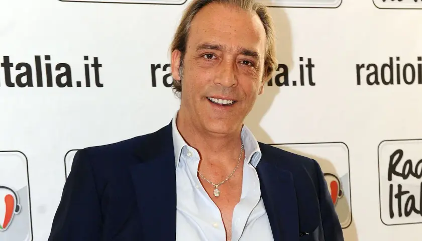 Luca Ward, noto per i suoi ruoli in film e programmi TV, sorride davanti a uno sfondo con il logo di Radio Italia.