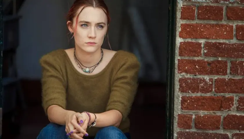 Ragazza pensierosa, con maglione verde e jeans, seduta vicino a un muro di mattoni rossi. Una scena che ricorda la recensione di Lady Bird.