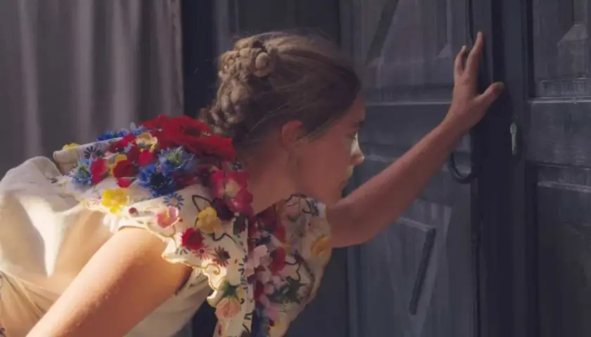 Una ragazza con un vestito fiorito, capelli raccolti, spinge una porta scura. Un'immagine che evoca la recensione di Midsommar.