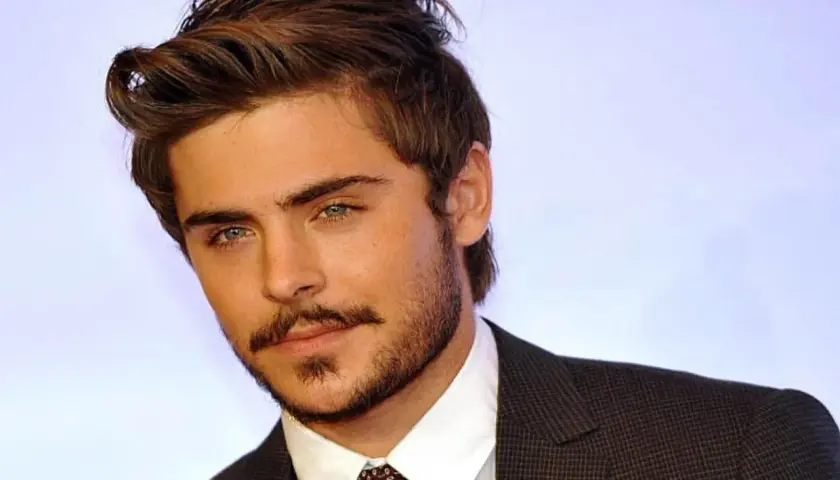 Primo piano di Zac Efron giovane, con sguardo intenso e barba curata, indossa una giacca scura.