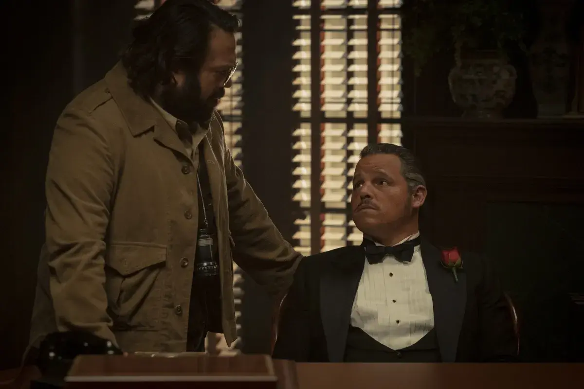 Un uomo con la barba e occhiali parla con un altro in smoking con una rosa. La scena suggerisce una recensione di un'offerta importante.
