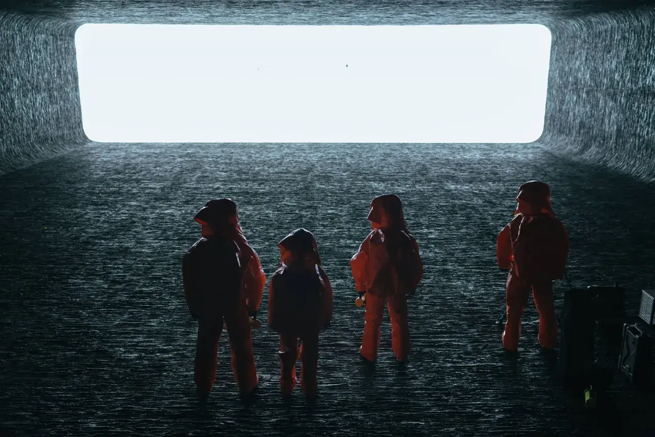 Persone in tute arancioni osservano un'enorme apertura luminosa, evocando scene di "Arrival" e la sua spiegazione di un evento alieno.