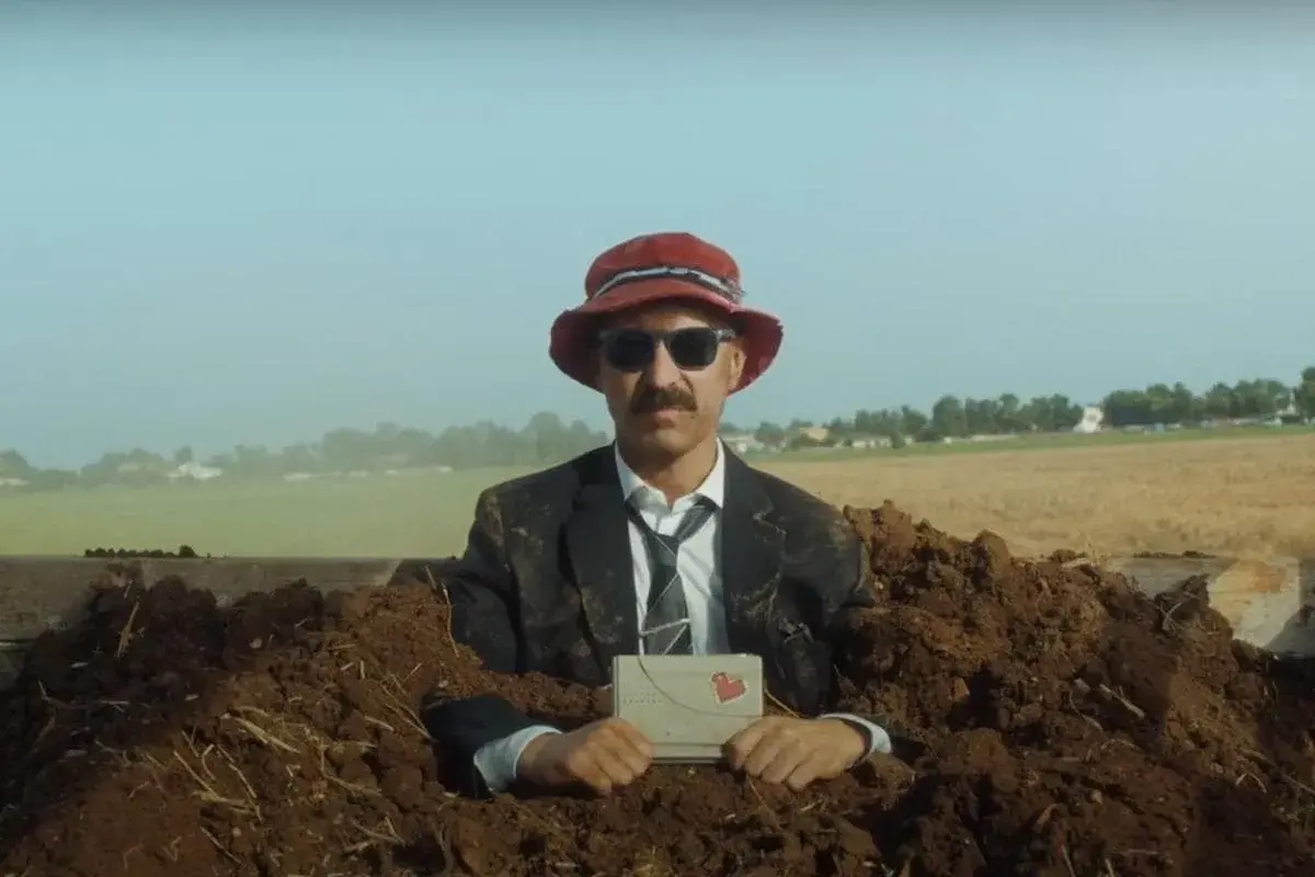 Uomo in abito e cappello rosso, sepolto nel terreno, tiene una lettera con un cuore. Una scena surreale per la recensione "il migliore dei mondi".