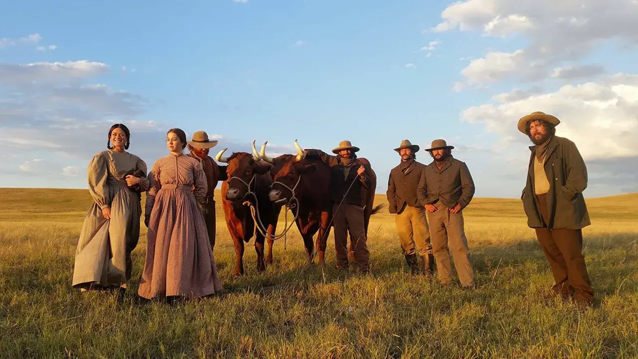 Un gruppo di persone in abiti d'epoca, con due buoi, in un campo dorato. Scena da "La ballata di Buster Scruggs", un film che merita una recensione.