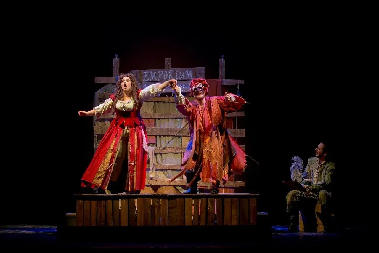 Scena teatrale con due attori in costume, uno con abito rosso e l'altra con maschera e vestiti arancioni. Sullo sfondo un cartello "Emporium". Le recensioni per Romeo è Giulietta lodano queste performance.